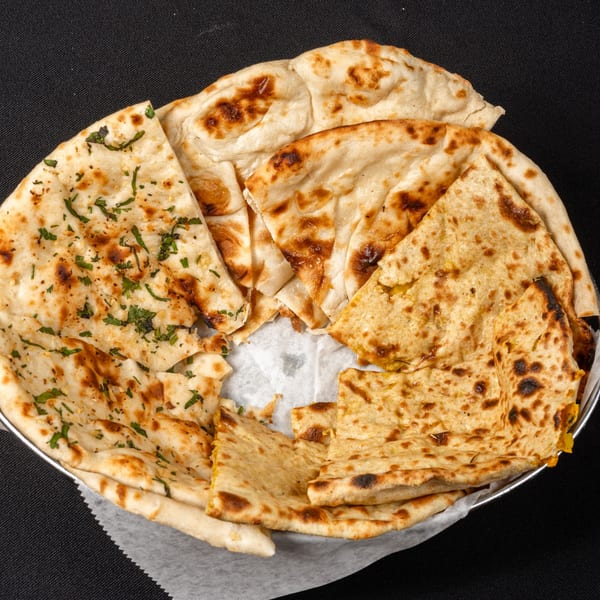 A basket of various Indian naan breads, including garlic naan and aloo paratha, on a black background.